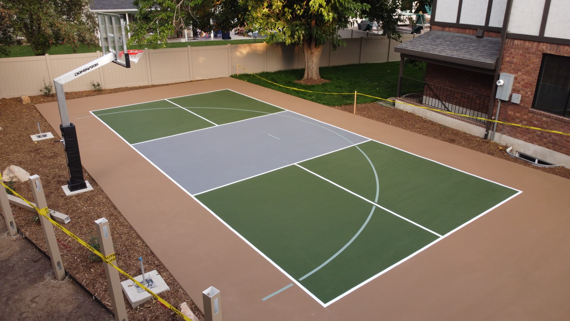 Outdoor basketball half-court with green and gray surface near a brick house and tree.
