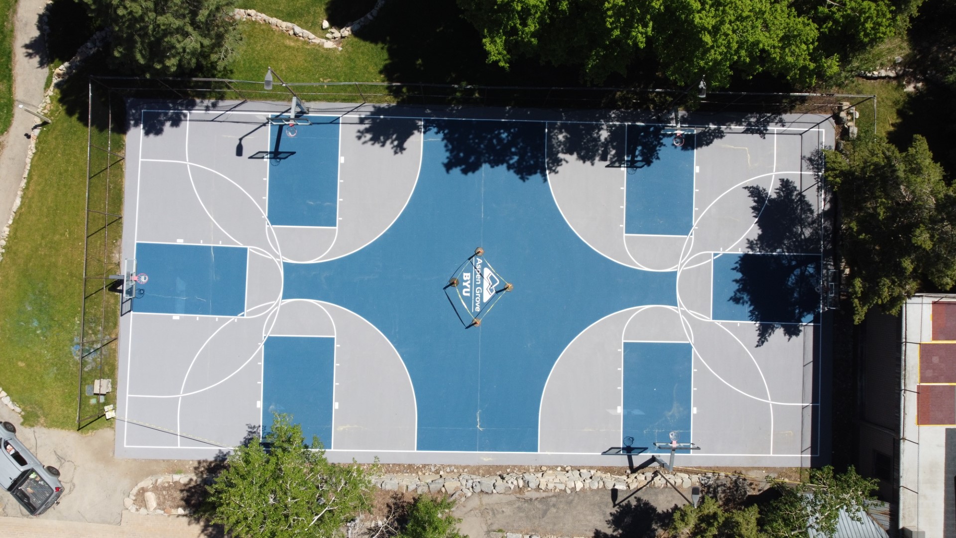 Aerial view of an outdoor basketball court with blue and gray paint and four hoops, surrounded by trees.