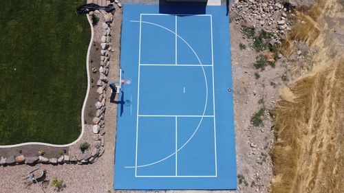 Aerial view of blue outdoor basketball court bordered by grass, rocks, and dry vegetation.