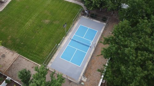 A fenced outdoor pickleball court with blue playing surfaces surrounded by green trees and grass.