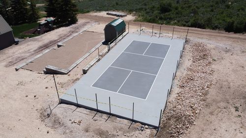 Newly constructed gray concrete pickleball court with white lines and a green-roofed shed nearby.