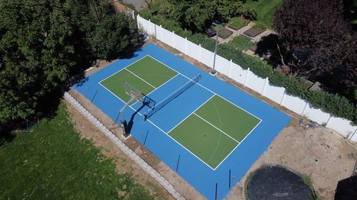 Backyard blue and green multipurpose sports court with basketball hoop, tennis net, and trampoline nearby.