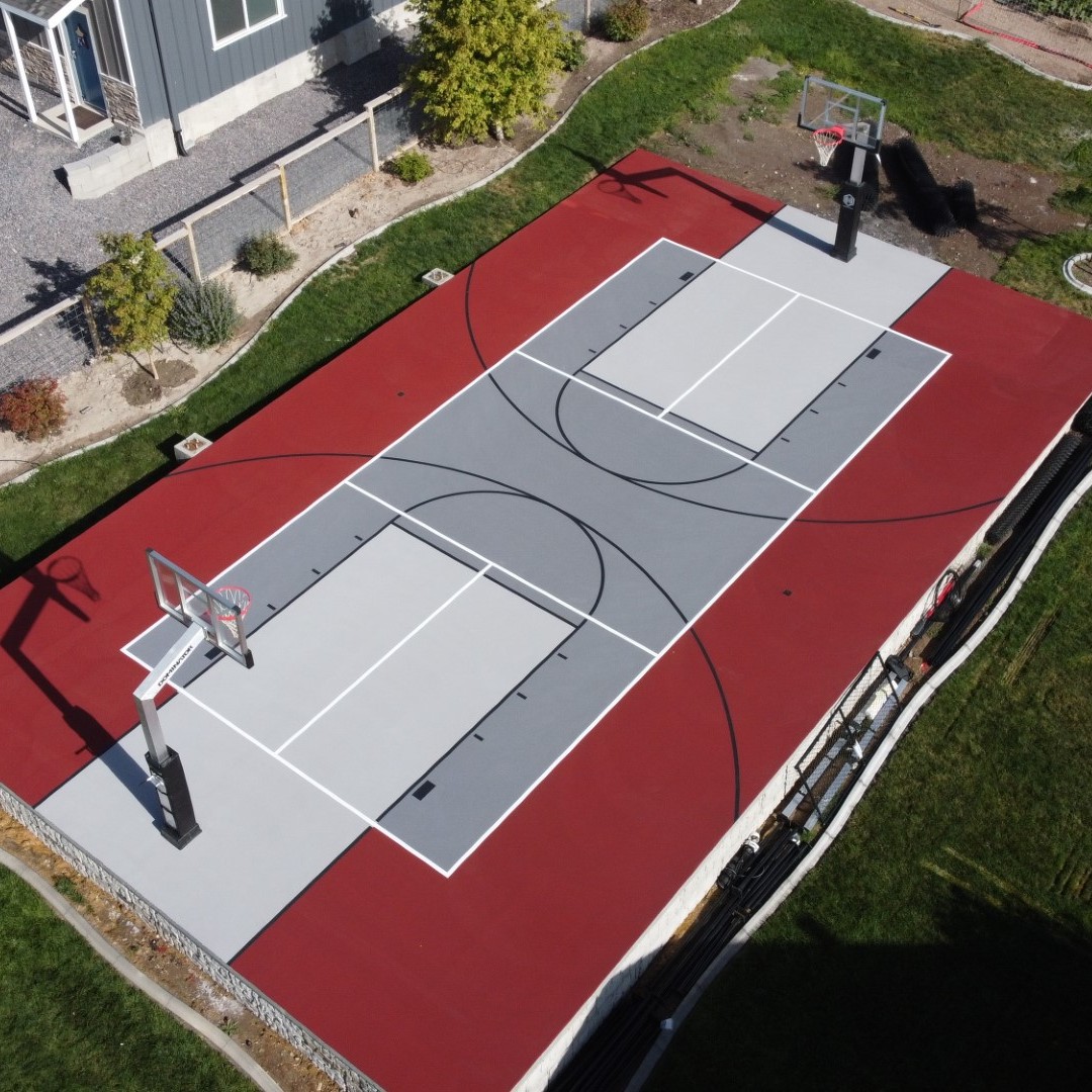 Aerial view of a backyard basketball court with a gray playing surface, red border, and two hoops beside a house.