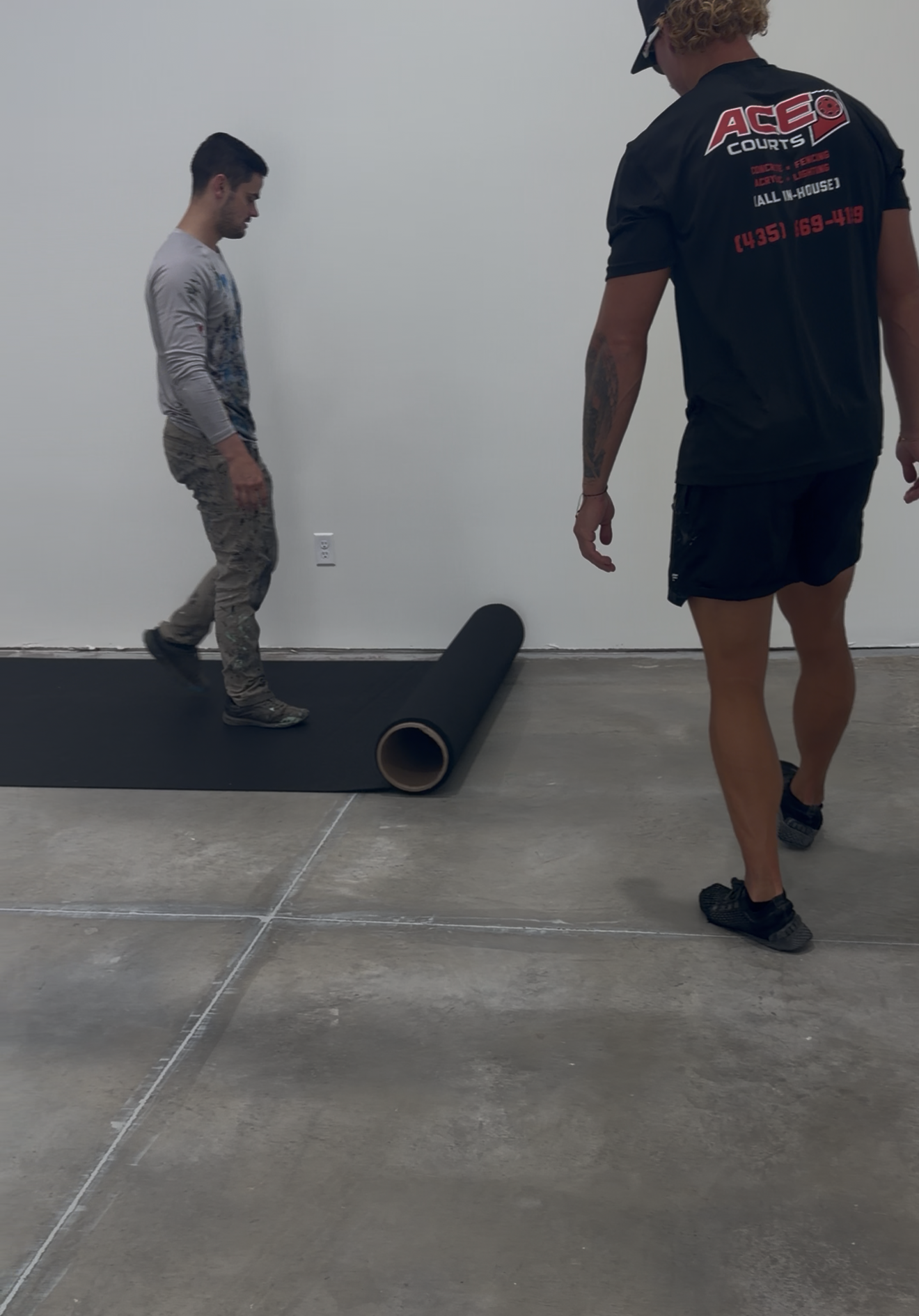 Two workers unrolling a dark mat over a concrete floor for an indoor court surface.