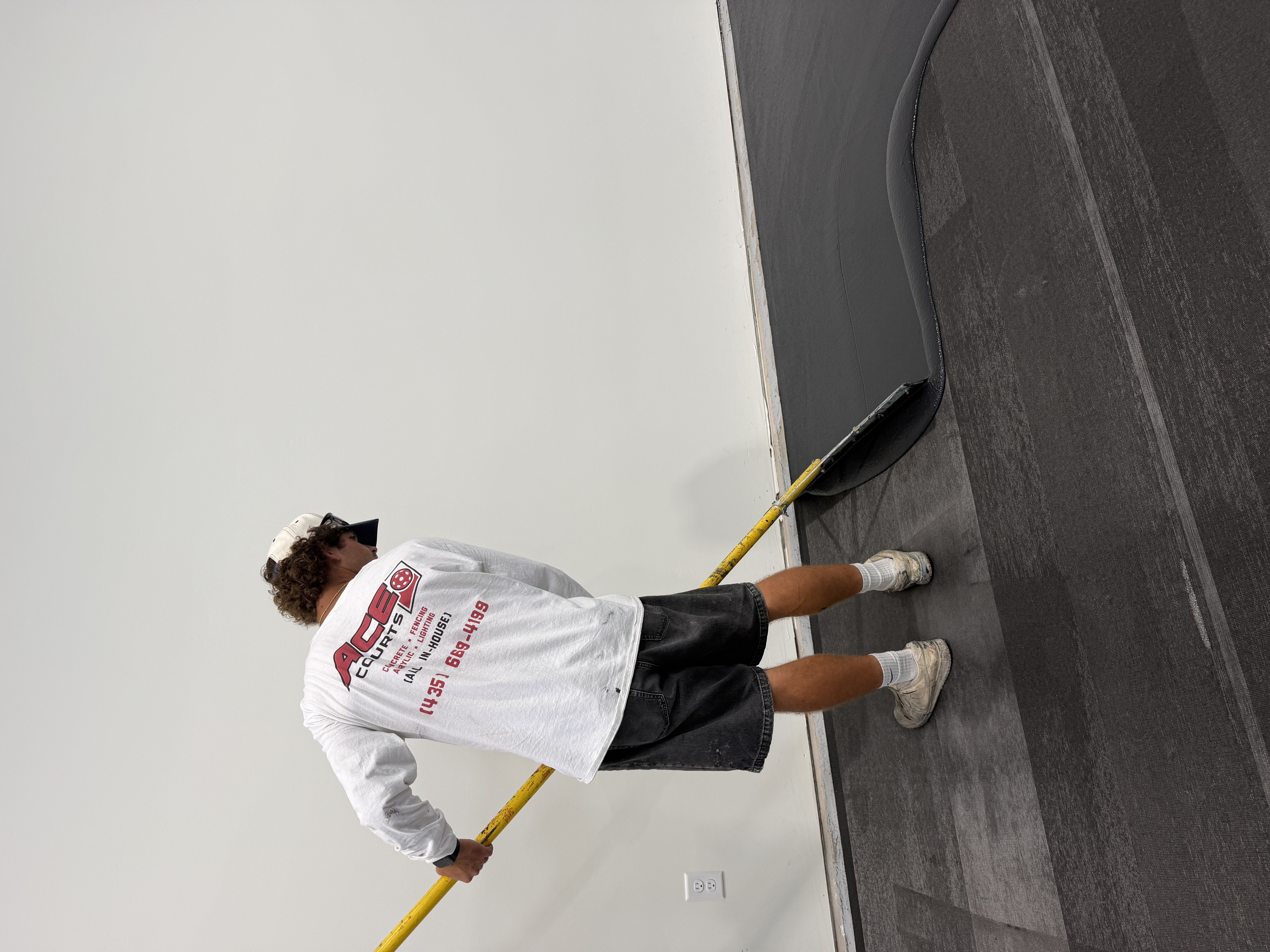 Worker spreading a dark surface coating across an indoor court with a long-handled squeegee.