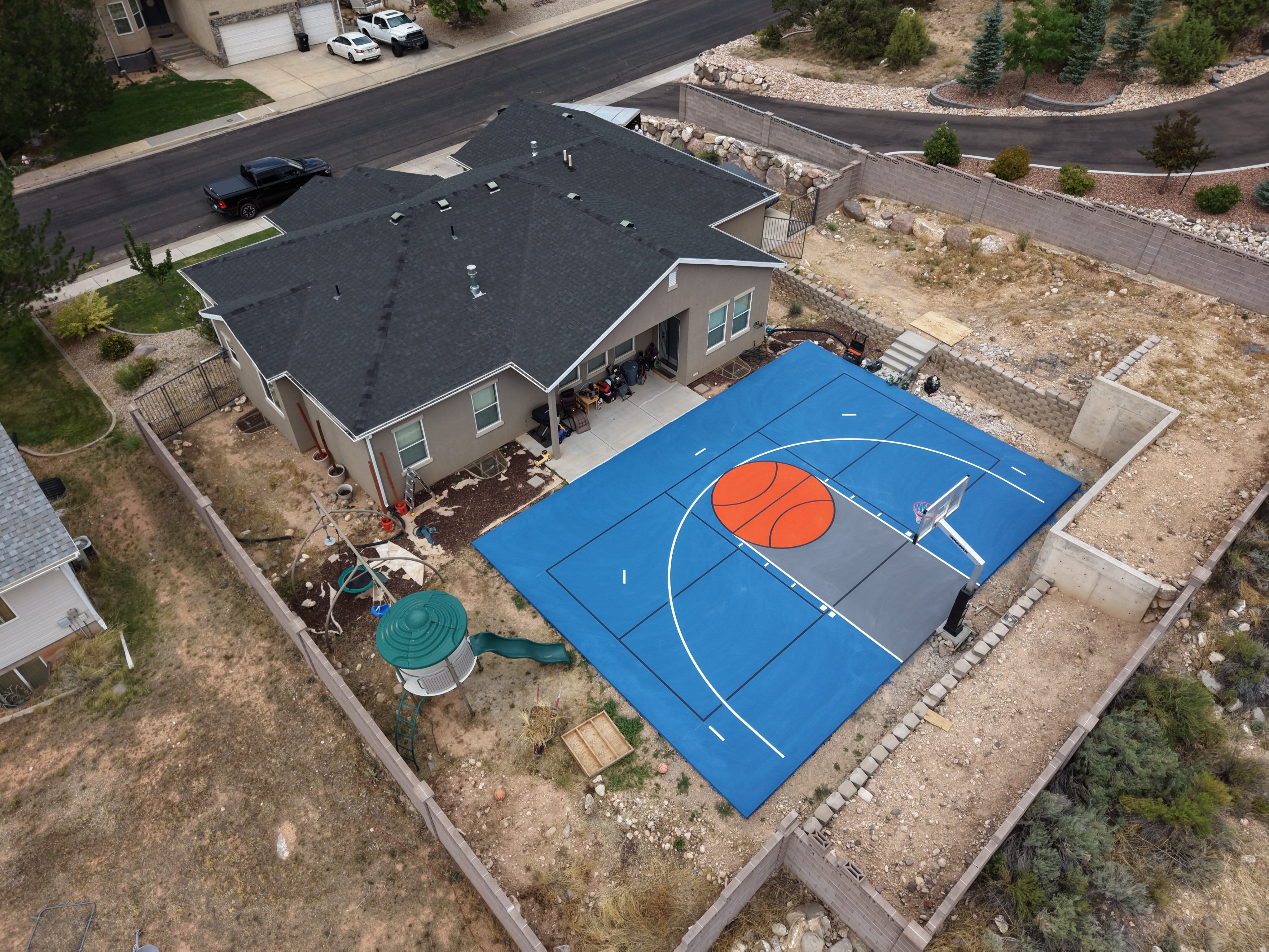 Aerial view of a blue backyard basketball court with an orange key, white lines, and a hoop beside a house.