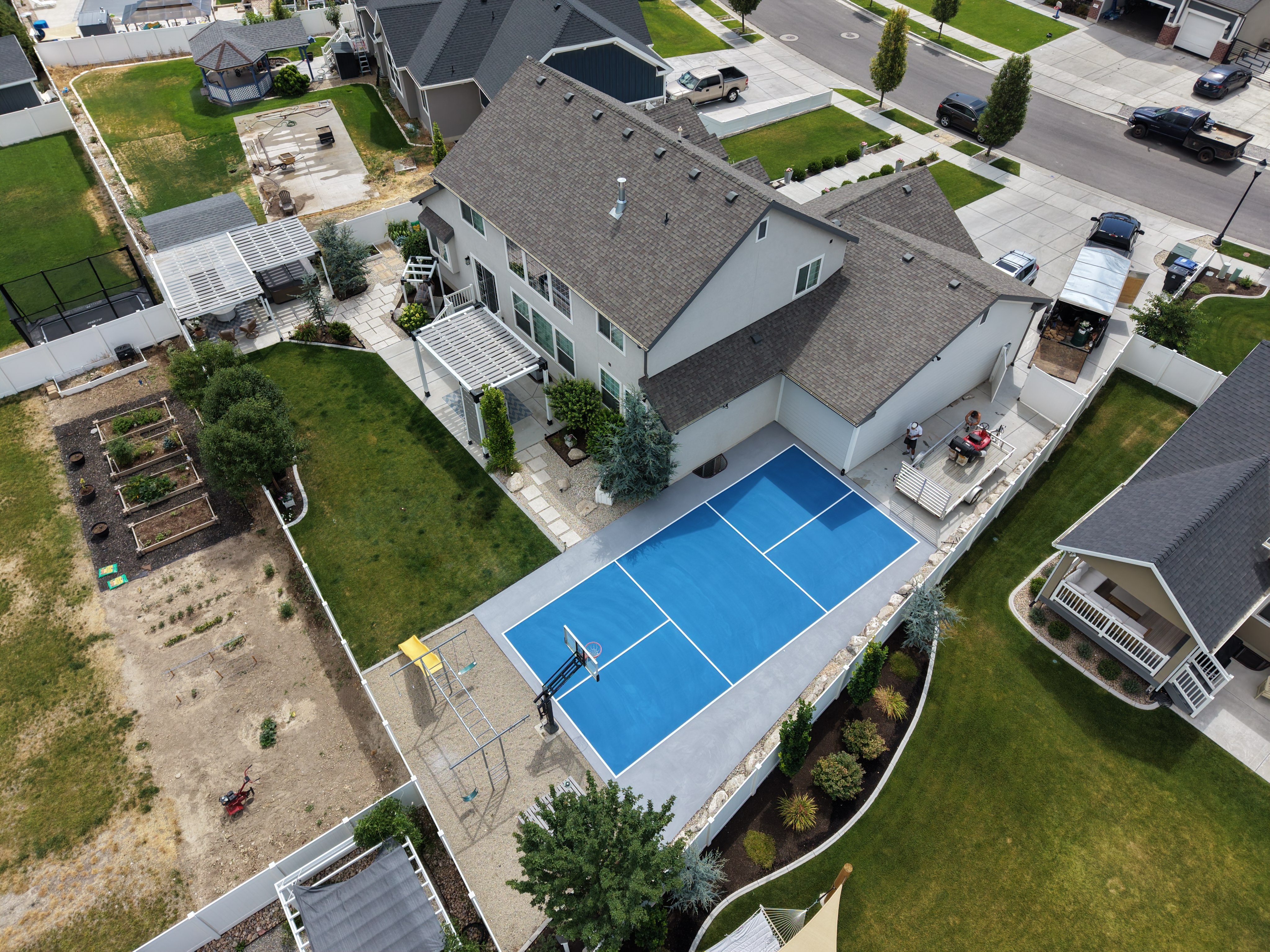 Aerial view of a blue pickleball court with a light perimeter beside a white house, bordered by lawn and trees.