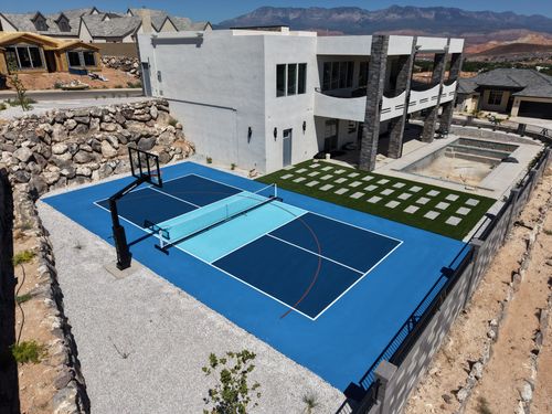 Blue pickleball court with a light blue center, white lines, and basketball hoop beside a modern home and checkerboard patio.