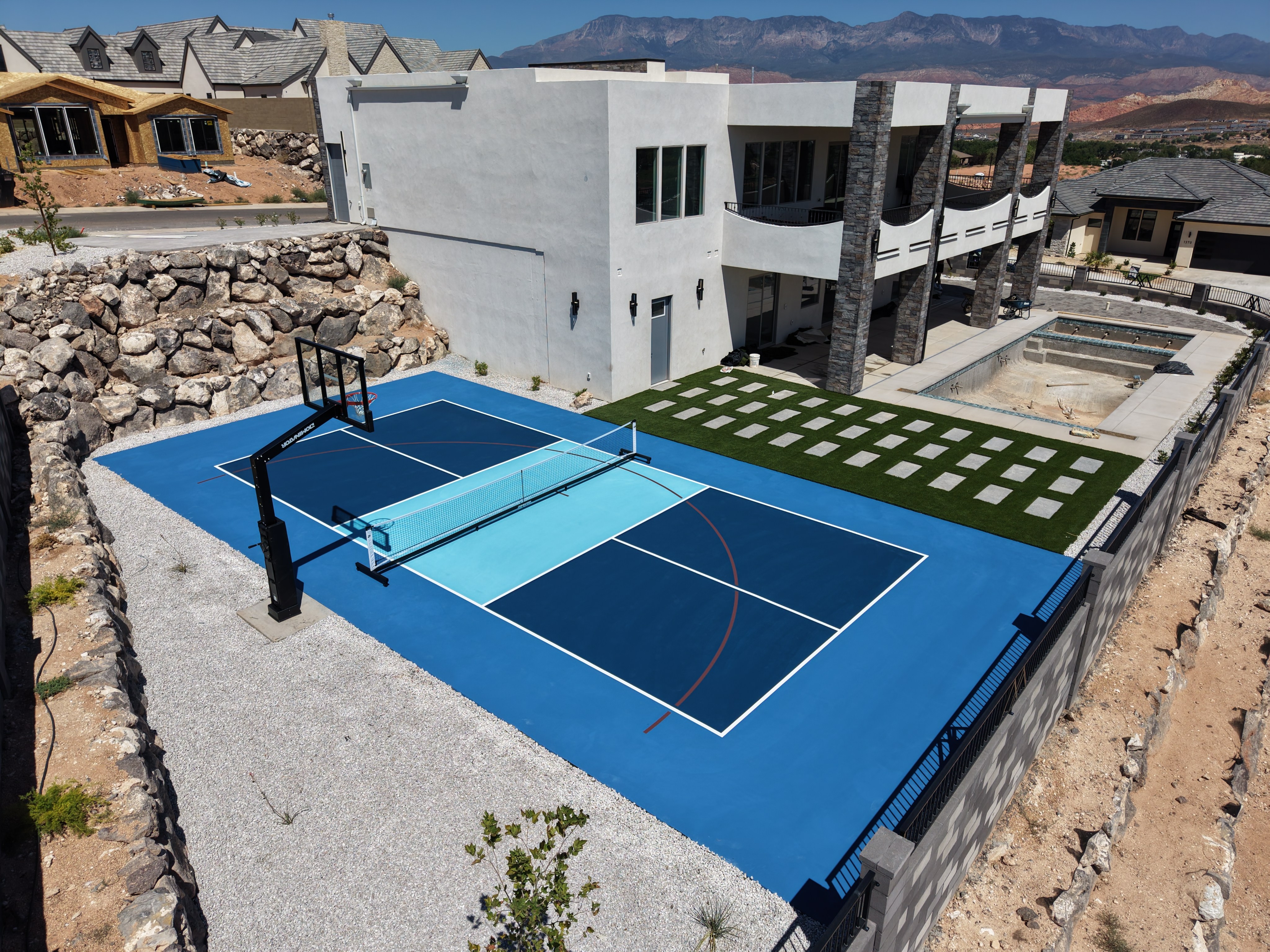 Blue pickleball court with a light blue center, white lines, and basketball hoop beside a modern home and checkerboard patio.