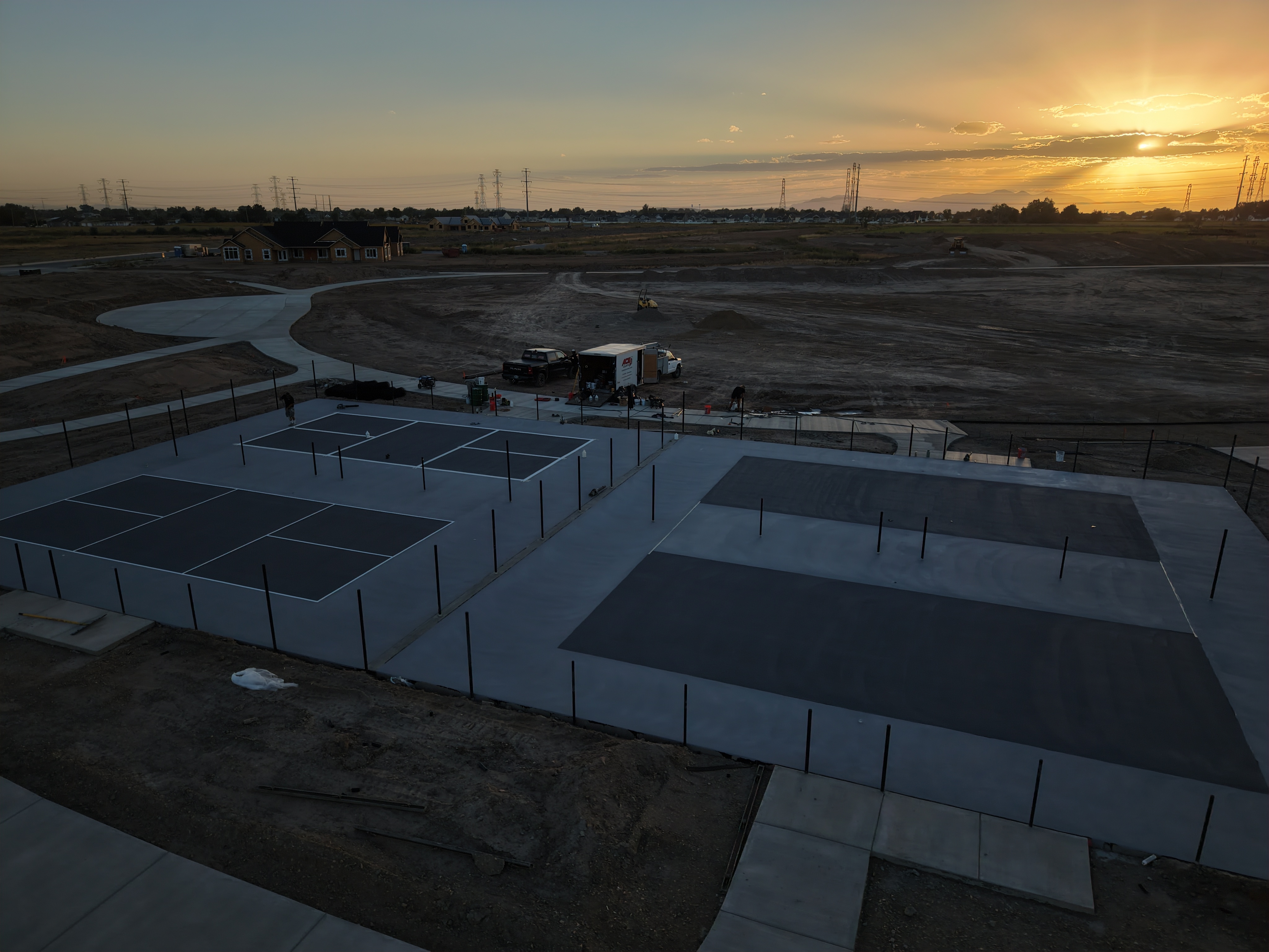 Fenced outdoor court complex at sunset, with multiple playing areas and paved walkways across an open site.