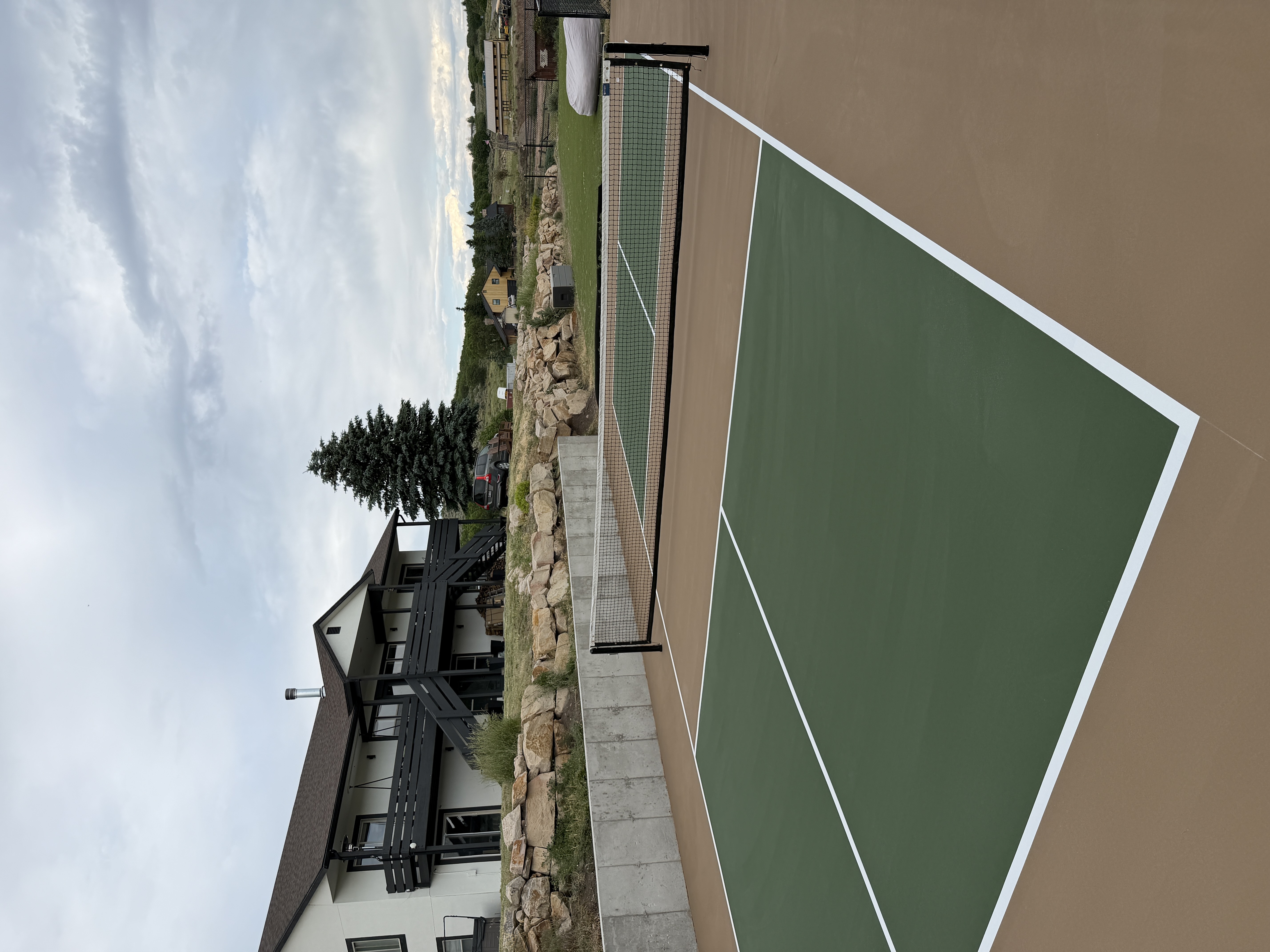 Green outdoor pickleball court with a tan perimeter and center net, beside a stone retaining wall and home.
