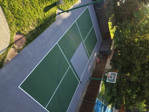 Green backyard pickleball court with a gray perimeter, center net, and basketball hoop, surrounded by trees.
