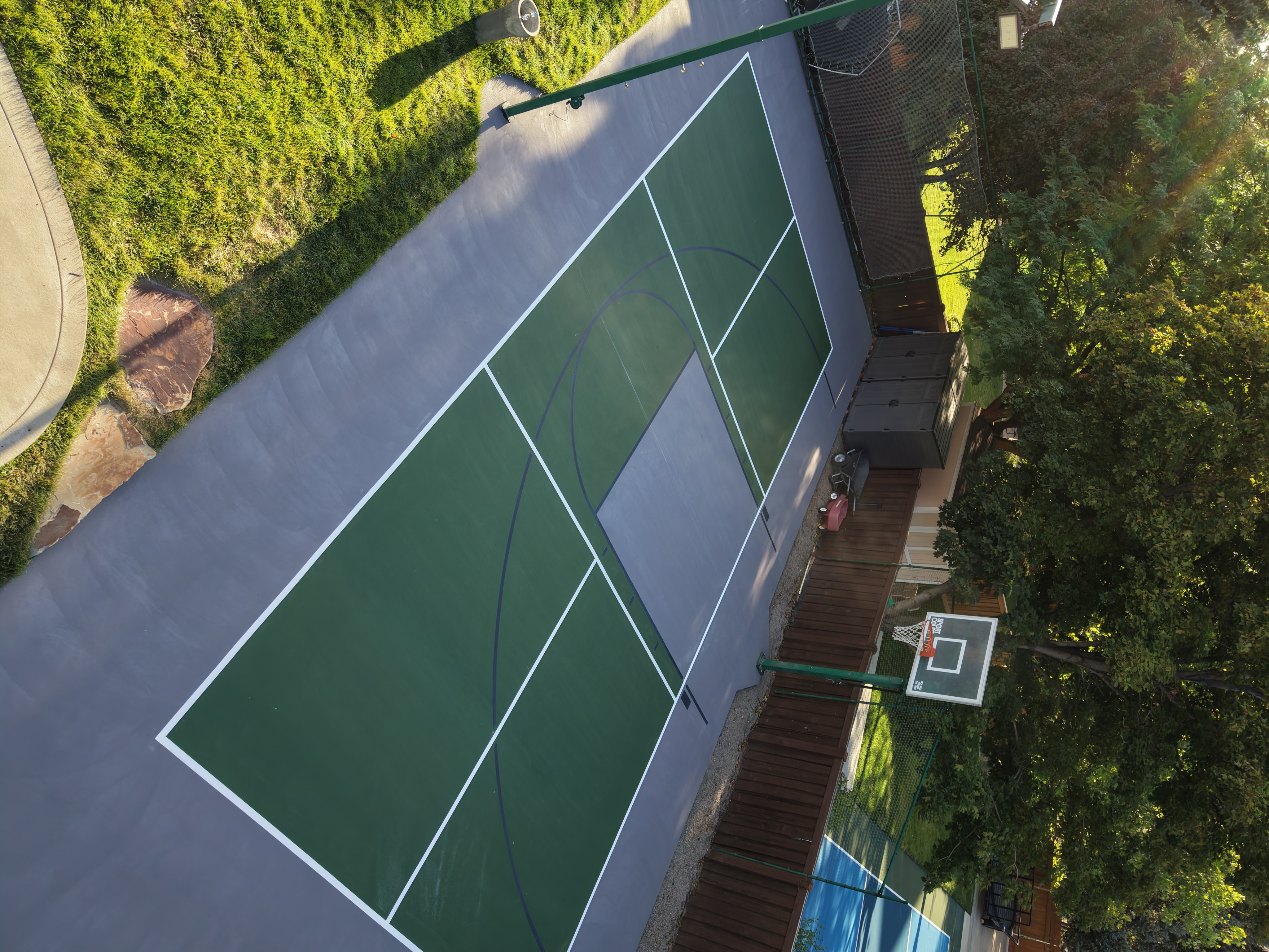 Green backyard pickleball court with a gray perimeter, center net, and basketball hoop, surrounded by trees.
