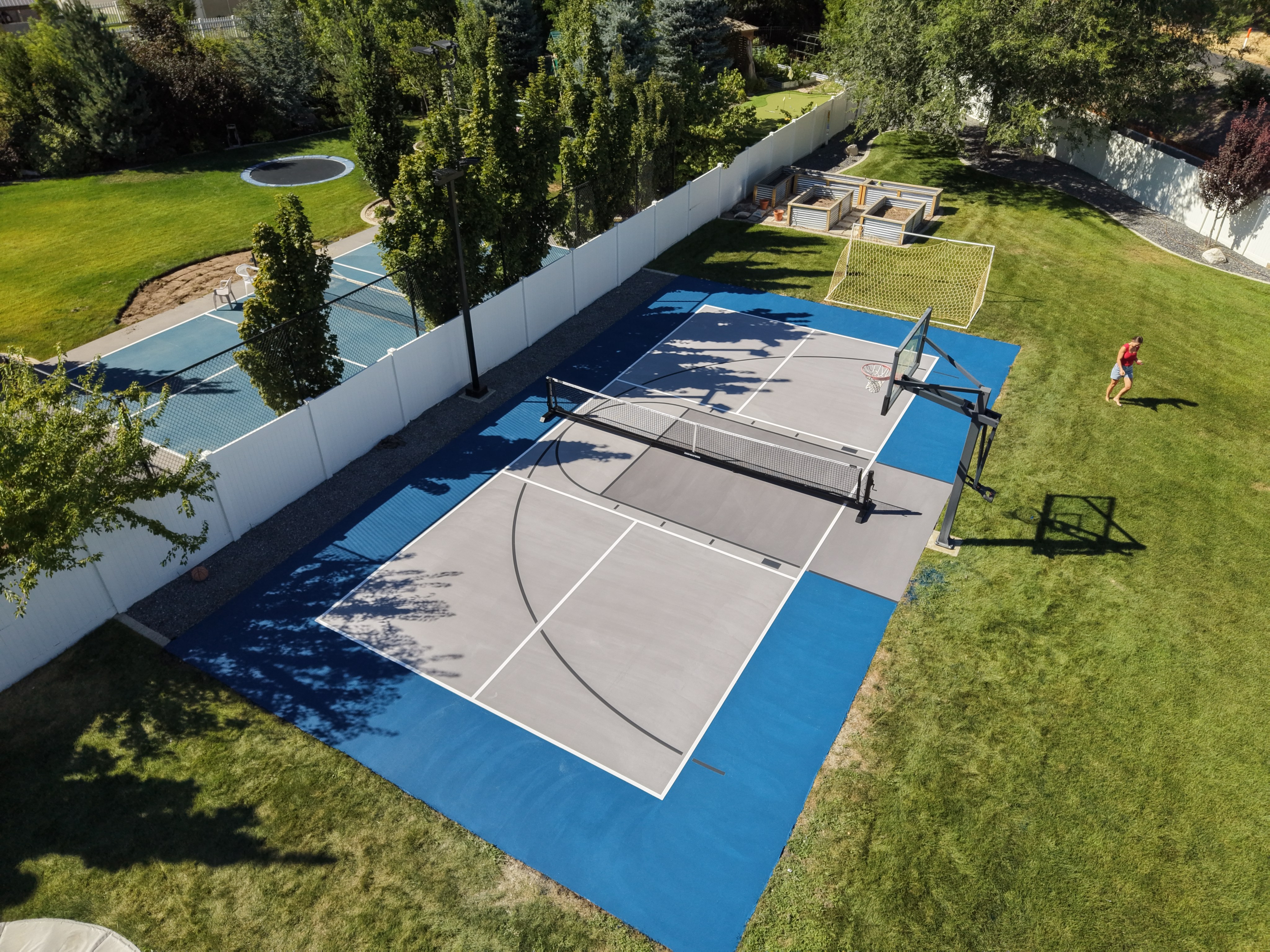 Backyard multi-sport court with a gray playing area, blue border, pickleball net, and basketball hoop.
