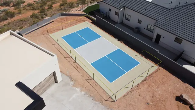 Blue outdoor net court with a light gray center and tan borders beside a home.