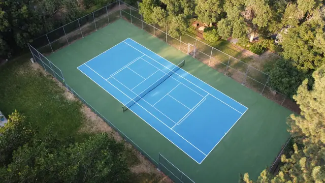 Blue and green fenced tennis court surrounded by mature trees.