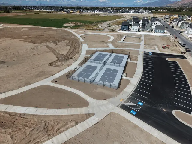 Aerial view of fenced gray outdoor courts with white game lines, paved walkways and adjacent parking.
