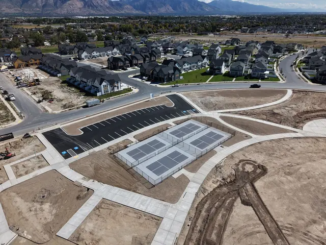Aerial view of fenced gray outdoor courts with white game lines, paved walkways and adjacent parking.