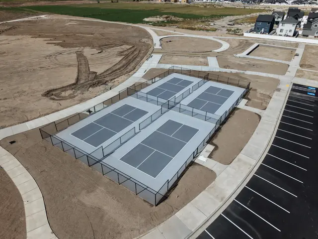 Aerial view of fenced gray outdoor courts with white game lines, paved walkways and adjacent parking.