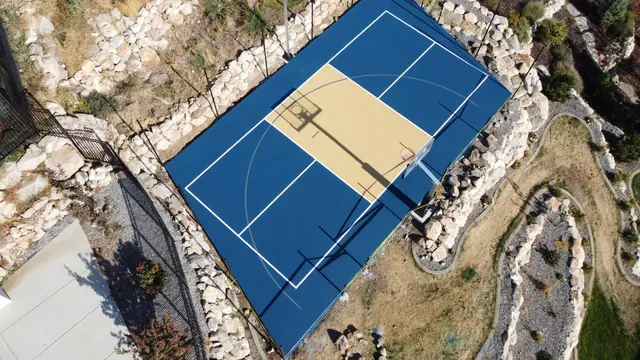 Blue and tan basketball court with black fencing and stone landscaping.