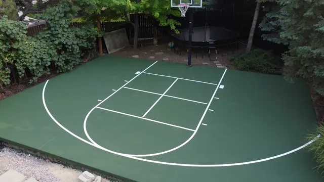 Green basketball half court with white markings surrounded by evergreen trees.