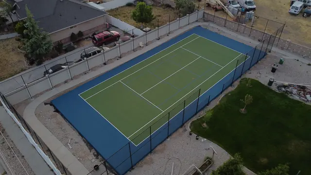 Green tennis playing surface with a blue perimeter, net and black fencing.