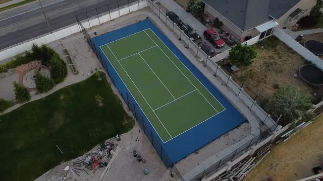 Green tennis playing surface with a blue perimeter, net and black fencing.
