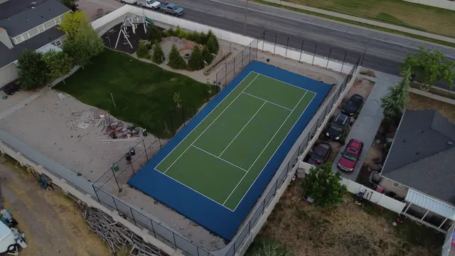 Green tennis playing surface with a blue perimeter, net and black fencing.