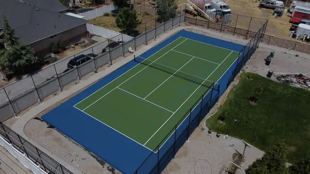 Green tennis playing surface with a blue perimeter, net and black fencing.