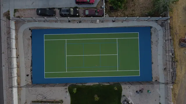 Green tennis playing surface with a blue perimeter, net and black fencing.