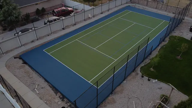 Green tennis playing surface with a blue perimeter, net and black fencing.