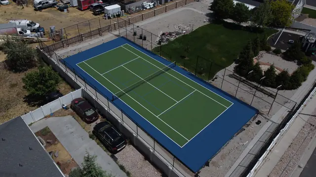 Green tennis playing surface with a blue perimeter, net and black fencing.