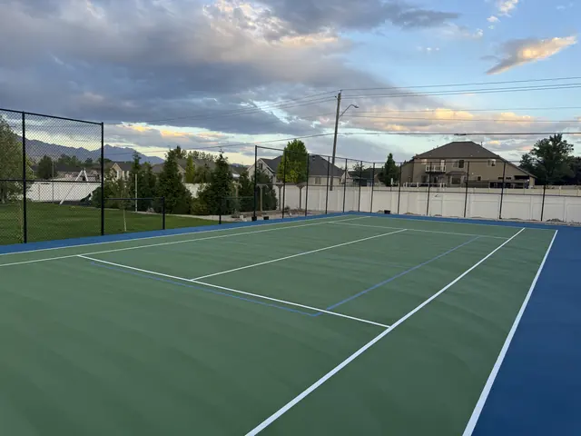 Green tennis playing surface with a blue perimeter, net and black fencing.