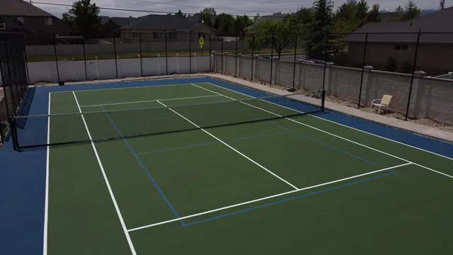 Green tennis playing surface with a blue perimeter, net and black fencing.