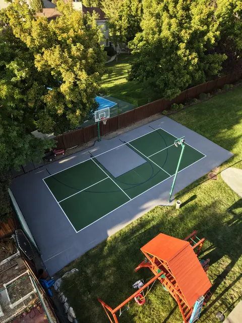 Green and gray backyard court with basketball and net-game markings beside a lawn.