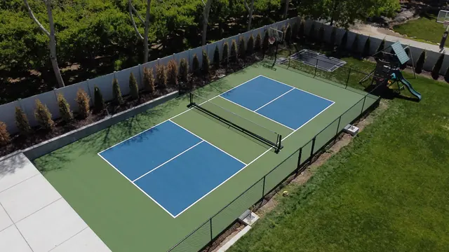 Blue pickleball playing areas with green borders, a center net and black perimeter fencing.
