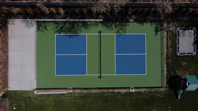 Blue pickleball playing areas with green borders, a center net and black perimeter fencing.