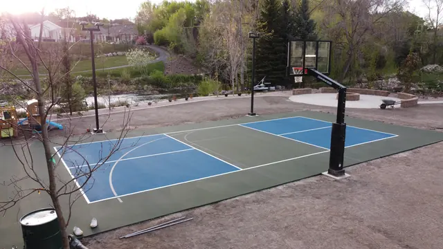 Blue and green backyard basketball court with two hoops and tall light poles.
