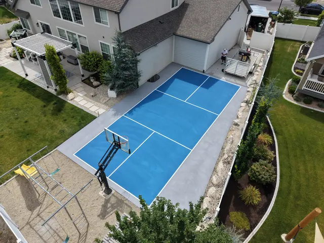 Blue backyard court with white net-game markings and a basketball hoop beside a curved lawn.