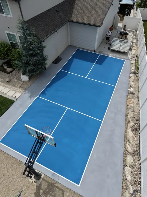 Blue backyard court with white net-game markings and a basketball hoop beside a curved lawn.