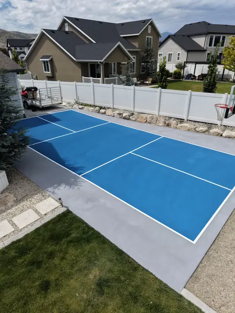 Blue backyard court with white net-game markings and a basketball hoop beside a curved lawn.