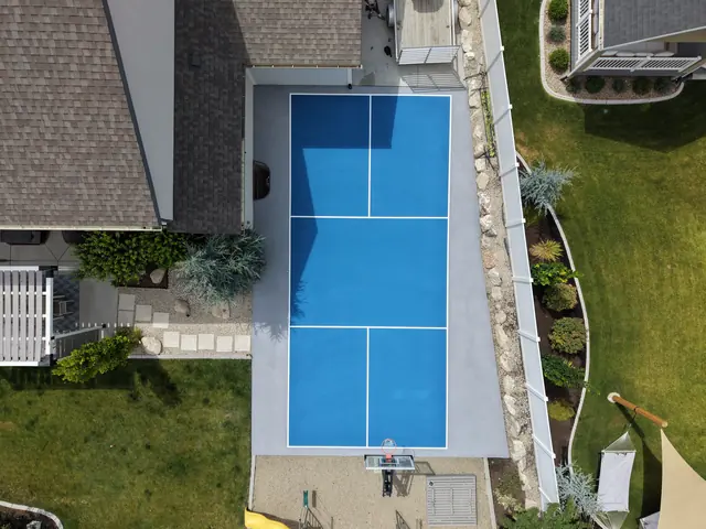Blue backyard court with white net-game markings and a basketball hoop beside a curved lawn.