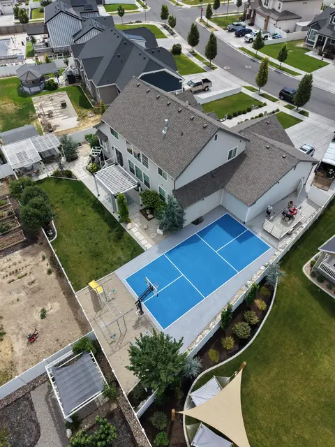 Blue backyard court with white net-game markings and a basketball hoop beside a curved lawn.