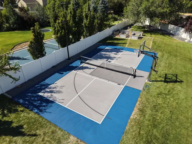 Gray backyard multi-sport court with blue borders, a net and basketball hoops beside a lawn.