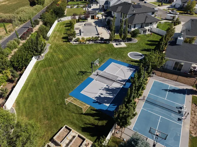 Gray backyard multi-sport court with blue borders, a net and basketball hoops beside a lawn.