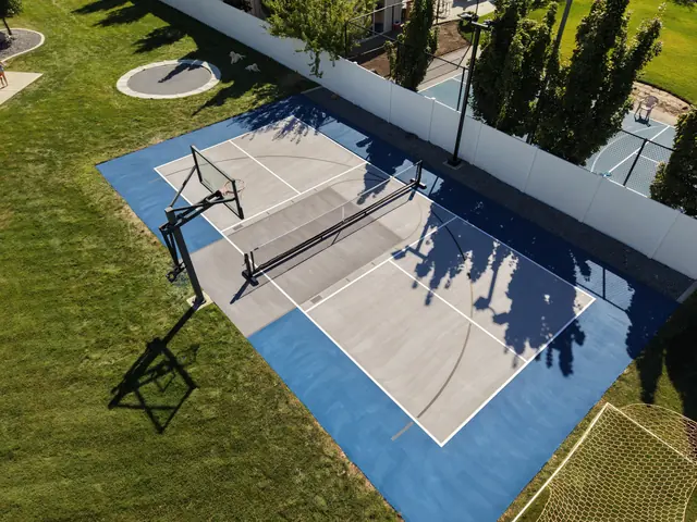 Gray backyard multi-sport court with blue borders, a net and basketball hoops beside a lawn.