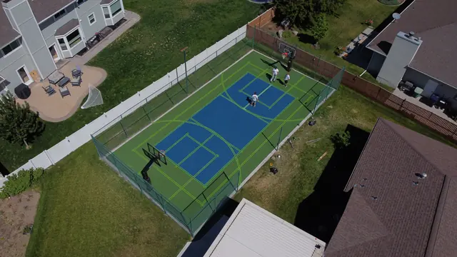 Blue and green backyard court with black fencing, a center net and basketball hoops.