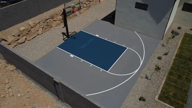 Compact gray basketball half court with a dark blue key beside a home.