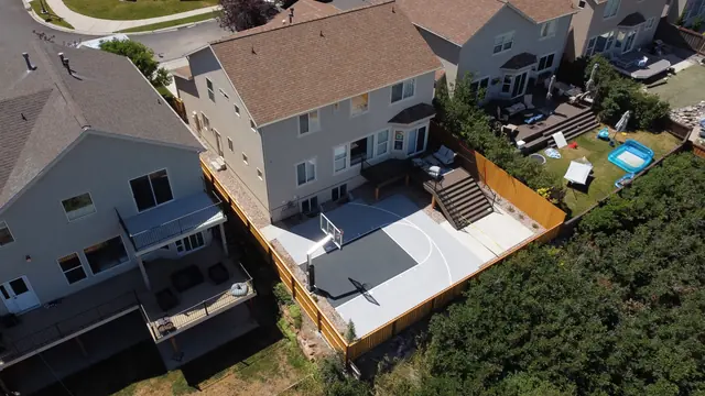 Gray backyard basketball half court with a dark key, white lines and wood perimeter fencing.