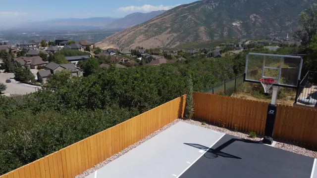 Gray backyard basketball half court with a dark key, white lines and wood perimeter fencing.